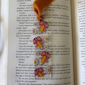 May include: A clear rectangular bookmark featuring a repeating pattern of yellow pencils with pink erasers, pink bows, and white daisies with yellow centers. The bookmark is topped with a frayed mustard yellow fabric ribbon tassel.