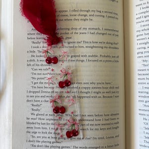 May include: A clear rectangular bookmark featuring a repeating pattern of red cherries with light pink bows and small star accents. The bookmark is topped with a frayed deep red fabric ribbon and is placed inside an open book by Jojo Moyes.