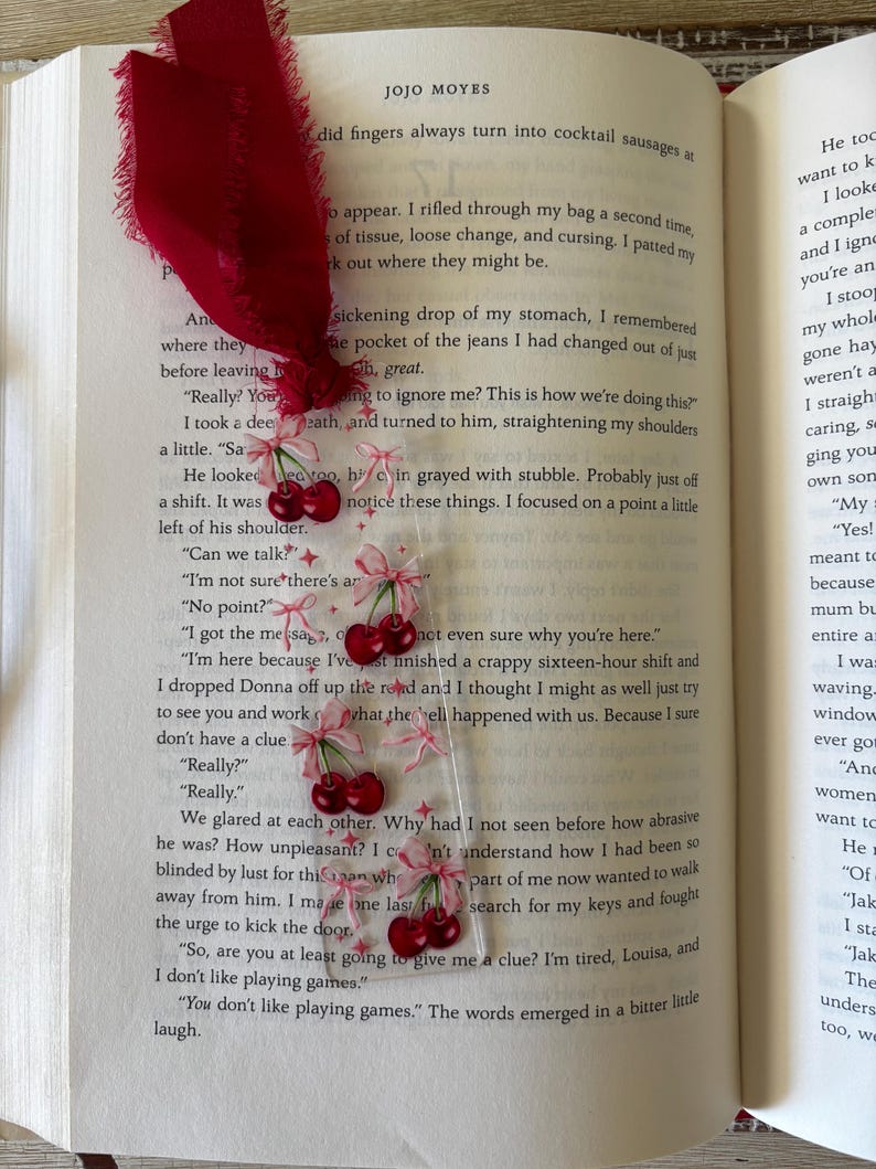 May include: A clear rectangular bookmark featuring a repeating pattern of red cherries with light pink bows and small star accents. The bookmark is topped with a frayed deep red fabric ribbon and is placed inside an open book by Jojo Moyes.