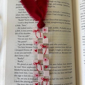 May include: A rectangular clear acrylic bookmark featuring a white and light pink checkerboard pattern. The design is decorated with small pink flowers, red hearts, and red ribbon bows. A frayed dark red fabric ribbon is tied at the top.