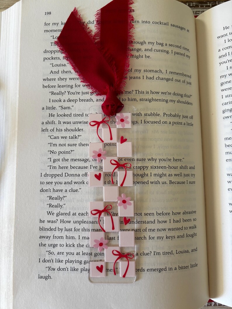 May include: A rectangular clear acrylic bookmark featuring a white and light pink checkerboard pattern. The design is decorated with small pink flowers, red hearts, and red ribbon bows. A frayed dark red fabric ribbon is tied at the top.