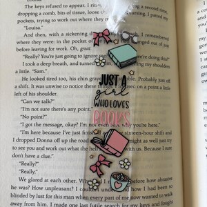 May include: A clear rectangular bookmark with a white ribbon topper, featuring the black and pink text "JUST A girl WHO LOVES BOOKS" surrounded by illustrations of a teal book, pink book, pink bows, white flowers, glasses, and a mug.