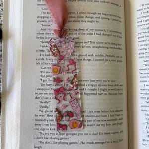 May include: A clear rectangular bookmark featuring a repeating pattern of pink bows, strawberries, coffee mugs, and tumblers with floral accents. The bookmark is topped with a frayed dusty rose fabric ribbon and is placed inside an open book.