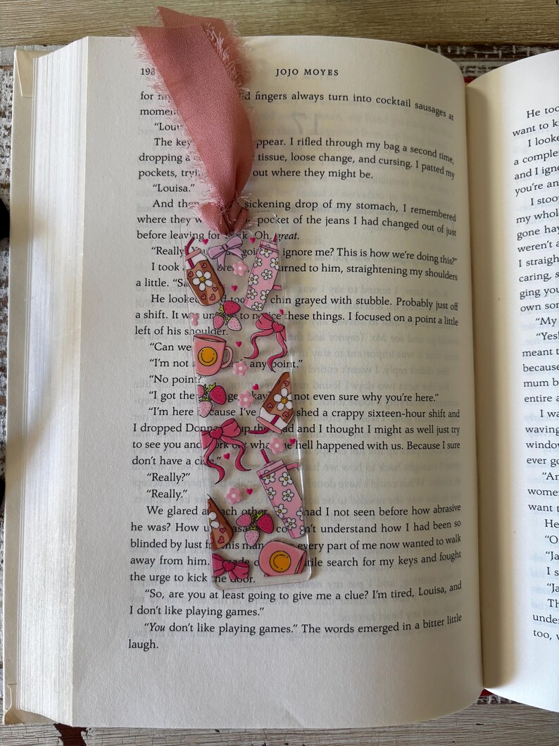 May include: A clear rectangular bookmark featuring a repeating pattern of pink bows, strawberries, coffee mugs, and tumblers with floral accents. The bookmark is topped with a frayed dusty rose fabric ribbon and is placed inside an open book.
