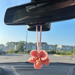 May include: A handmade, pink crocheted flower car charm hanging from a car's rearview mirror. The flower has a yellow center and is suspended by a matching pink cord. The background shows a sunny outdoor scene.