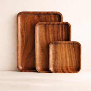 Puede incluir: Juego de tres bandejas de madera de diferentes tamaños. Las bandejas son rectangulares con esquinas redondeadas y un acabado natural de vetas de madera. La bandeja más grande mide unos 40 cm de largo, la mediana 30 cm y la más pequeña 20 cm.