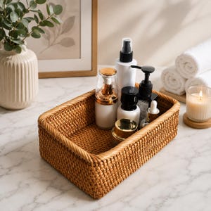 May include: A rectangular woven rattan storage basket containing several skincare bottles and jars. The basket features a tight, natural-colored weave with a braided rim. Inside are various containers, including a white spray bottle, a black pump bottle, and small glass jars with gold and black lids.