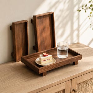 Puede incluir: Tres bandejas de madera marrón oscuro. Una bandeja contiene una porción de pastel con una fresa y un vaso de agua. Las bandejas son rectangulares con bordes elevados y se apoyan en pequeños pies cuadrados. La veta de la madera es visible.