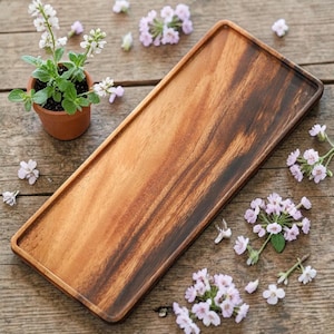 Könnte beinhalten: Ein rechteckiges Serviertablett aus Holz mit erhöhtem Rand, das ein reichhaltiges braunes Maserungsmuster aufweist. Eine kleine Topfpflanze mit weißen Blüten und verstreuten rosa Blüten schmückt die Holzoberfläche und schafft eine natürliche, rustikale Ästhetik.