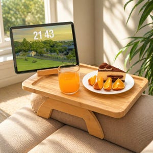 Könnte beinhalten: Ein Laptop-Tisch aus Bambus mit einem Tablet, das die Uhrzeit 21:43 anzeigt, einem Glas Orangensaft, einem Teller mit einem Stück Kuchen und Orangenspalten. Der Tisch liegt auf einem beigen Sofa.