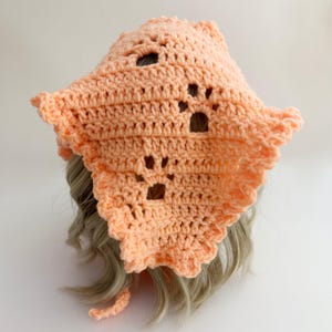 May include: A peach-colored, crocheted hat with paw print cutouts. The hat has a scalloped edge and a small tie. The hat is shown on a mannequin head with light blonde hair. The hat is handmade.