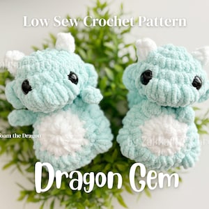 May include: Two crocheted dragon toys in seafoam green and white. The dragons have black eyes and white horns. The text "Low Sew Crochet Pattern" and "Dragon Gem" are displayed. The dragon toys are sitting on a bed of green leaves.
