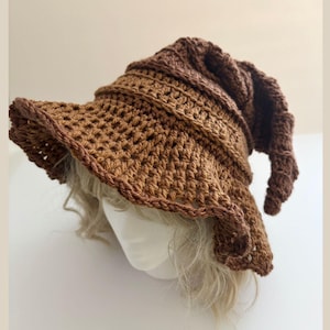 May include: A brown crochet hat with a wide brim and a pointed, curled tip. The hat is made of a textured, open-weave stitch and has a darker brown trim. The hat is designed to be worn on the head and is likely handmade.