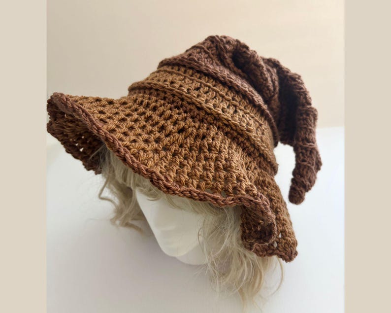 May include: A brown crochet hat with a wide brim and a pointed, curled tip. The hat is made of a textured, open-weave stitch and has a darker brown trim. The hat is designed to be worn on the head and is likely handmade.