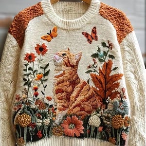 以下が含まれることがあります： オレンジ色のトラ猫、蝶、花が刺繍されたクリーム色のセーター。肩にはオレンジ色のアクセントがあり、裾と袖口はリブ編みです。猫は上を見上げており、カラフルな花のディテールに囲まれています。