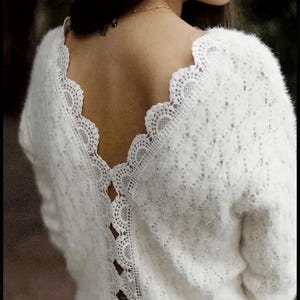 Könnte beinhalten: Ein weißer Langarmpullover mit zarter Spitzenborte am Ausschnitt und einem dekorativen Spitzeneinsatz am Rücken. Der Pullover hat ein weiches, strukturiertes Aussehen und einen gewellten Spitzenrand.