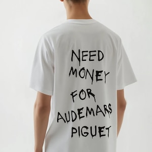 May include: White t-shirt with the text "NEED MONEY FOR AUDEMARS PIGUET" in black, handwritten-style lettering across the back. The shirt is short-sleeved and appears to be made of cotton.