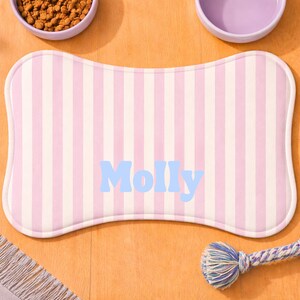Może przedstawiać: Mata na karmę dla zwierząt w kształcie kości z pionowymi r&oacute;żowymi i białymi paskami. Imię "Molly" jest wydrukowane na jasnoniebiesko. Widoczne są r&oacute;wnież dwie fioletowe miski i zabawka ze sznurka.
