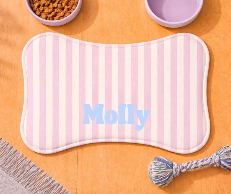 Może przedstawiać: Mata na karmę dla zwierząt w kształcie kości z pionowymi r&oacute;żowymi i białymi paskami. Imię "Molly" jest wydrukowane na jasnoniebiesko. Widoczne są r&oacute;wnież dwie fioletowe miski i zabawka ze sznurka.