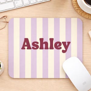 Könnte beinhalten: Rechteckiges Mauspad mit einem gestreiften Muster in Helllila und Creme. Der Name "Ashley" ist in einer kräftigen, kastanienbraunen Schriftart in der Mitte aufgedruckt. Eine weiße Computermaus befindet sich auf der rechten Seite des Mauspads.