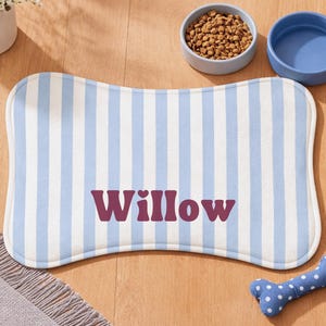 Może przedstawiać: Mata na karmę dla zwierząt w kształcie kości z niebiesko-białymi pionowymi paskami i imieniem "Willow" w kolorze bordowym. Obok maty znajdują się dwie ceramiczne miski, jedna z karmą dla ps&oacute;w, a druga pusta. Obecna jest r&oacute;wnież niebieska zabawka w kształcie kości w białe grochy.