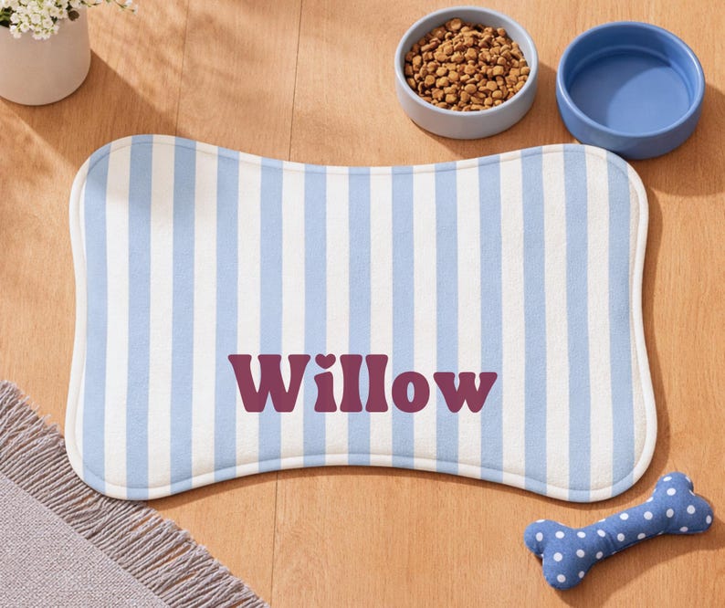 Może przedstawiać: Mata na karmę dla zwierząt w kształcie kości z niebiesko-białymi pionowymi paskami i imieniem "Willow" w kolorze bordowym. Obok maty znajdują się dwie ceramiczne miski, jedna z karmą dla ps&oacute;w, a druga pusta. Obecna jest r&oacute;wnież niebieska zabawka w kształcie kości w białe grochy.