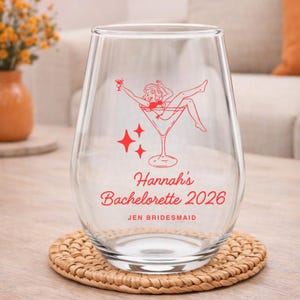 Op de afbeelding: Een helder, stuurloos wijnglas met een rode afbeelding van een vrouw in een martini-glas, met de tekst "Hannah's Bachelorette 2026" en "Jen Bridesmaid". Het glas staat op een ronde, geweven onderzetter.