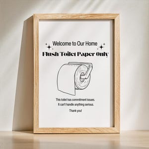 Peut inclure: Un cadre en bois clair encadre une impression avec un fond blanc. Le texte "Welcome to Our Home" et "Flush Toilet Paper Only" sont affichés. Un dessin d'un rouleau de papier toilette est en dessous. Le texte dit "This toilet has commitment issues."