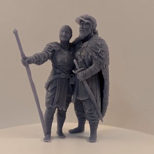 May include: A gray resin figurine of two figures, a man and a woman, in medieval attire. The man wears a hat, fur-lined cape, and carries a sword. The woman wears armor and holds a staff. The figures are posed together.