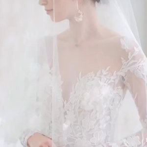 May include: A bride wearing a white lace wedding dress with long sleeves and a veil. She is holding her hands together in front of her.