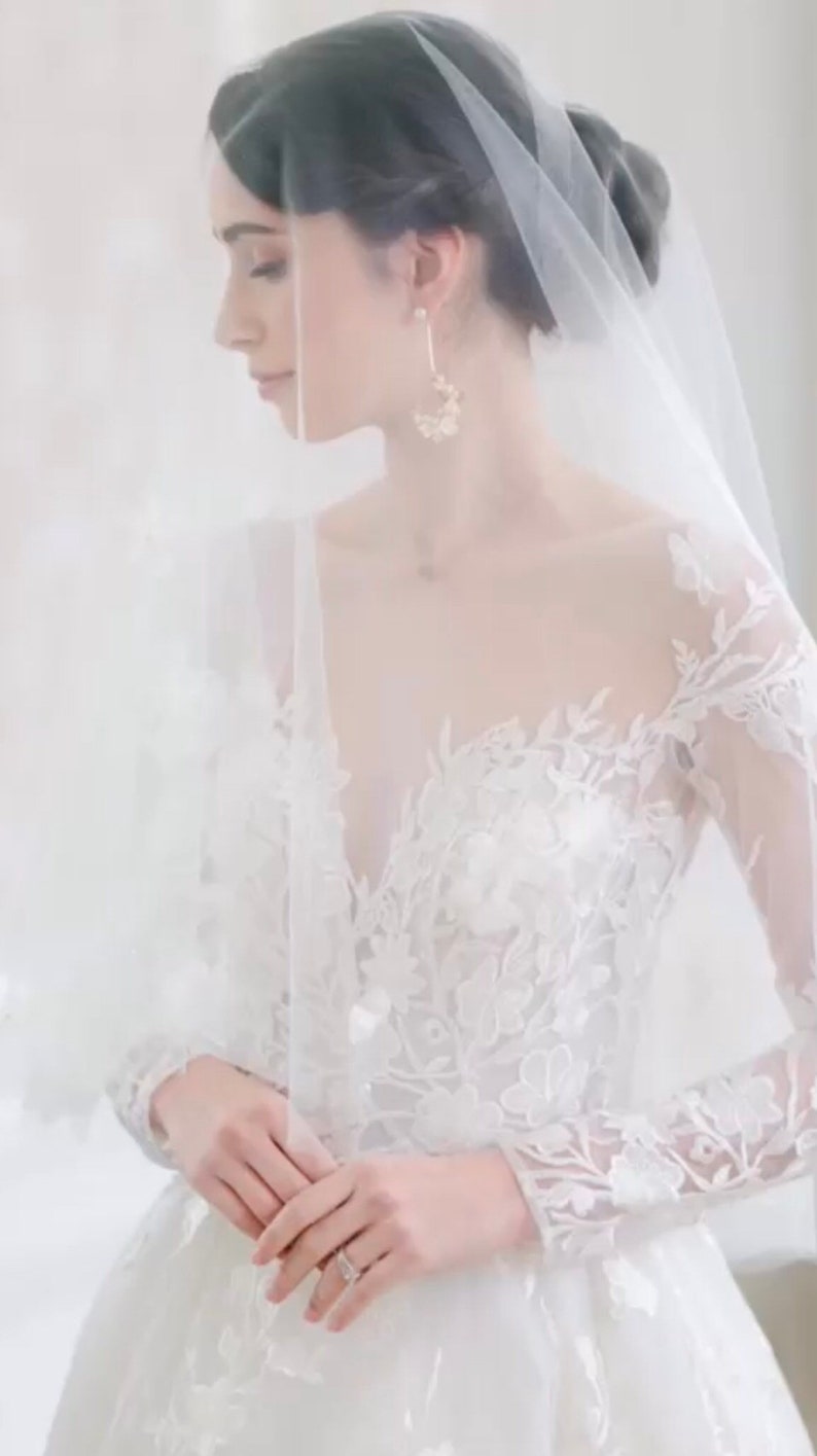 May include: A bride wearing a white lace wedding dress with long sleeves and a veil. She is holding her hands together in front of her.