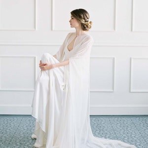 May include: A white wedding dress with a sheer cape and a long train. The dress is made of a soft, flowing fabric and has a simple, elegant design.