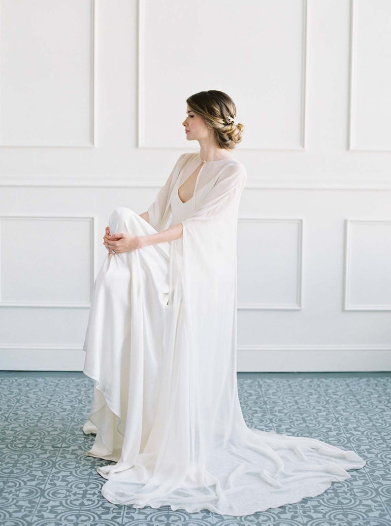 May include: A white wedding dress with a sheer cape and a long train. The dress is made of a soft, flowing fabric and has a simple, elegant design.
