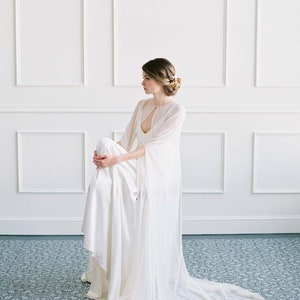 May include: A woman wearing a white wedding dress with a sheer cape. The dress has a low neckline and a fitted bodice. The cape is long and flowing, and it has a keyhole opening at the neckline. The woman is sitting on the floor, and her hair is styled in a bun. The background is a white wall with white trim.