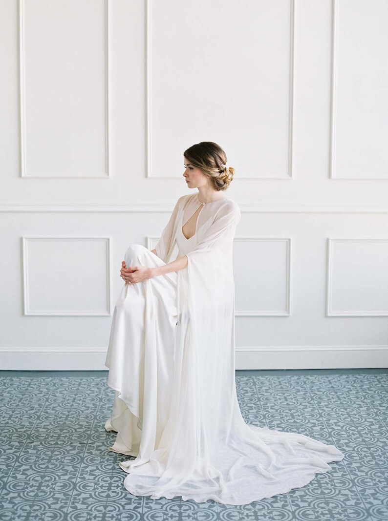 May include: A woman wearing a white wedding dress with a sheer cape. The dress has a low neckline and a fitted bodice. The cape is long and flowing, and it has a keyhole opening at the neckline. The woman is sitting on the floor, and her hair is styled in a bun. The background is a white wall with white trim.