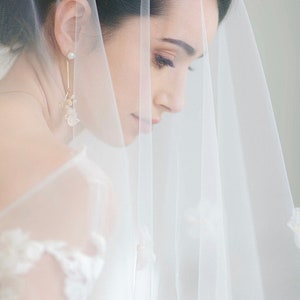 May include: A bride wearing a white veil with delicate floral accents. The veil is sheer and covers her face, with only her eyes and part of her nose visible. She is wearing a pair of gold earrings with pearl accents.