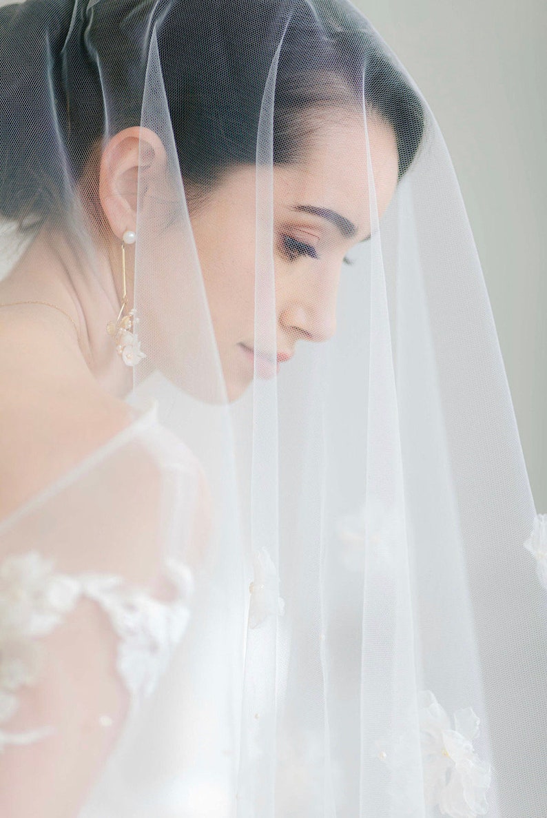 May include: A bride wearing a white veil with delicate floral accents. The veil is sheer and covers her face, with only her eyes and part of her nose visible. She is wearing a pair of gold earrings with pearl accents.
