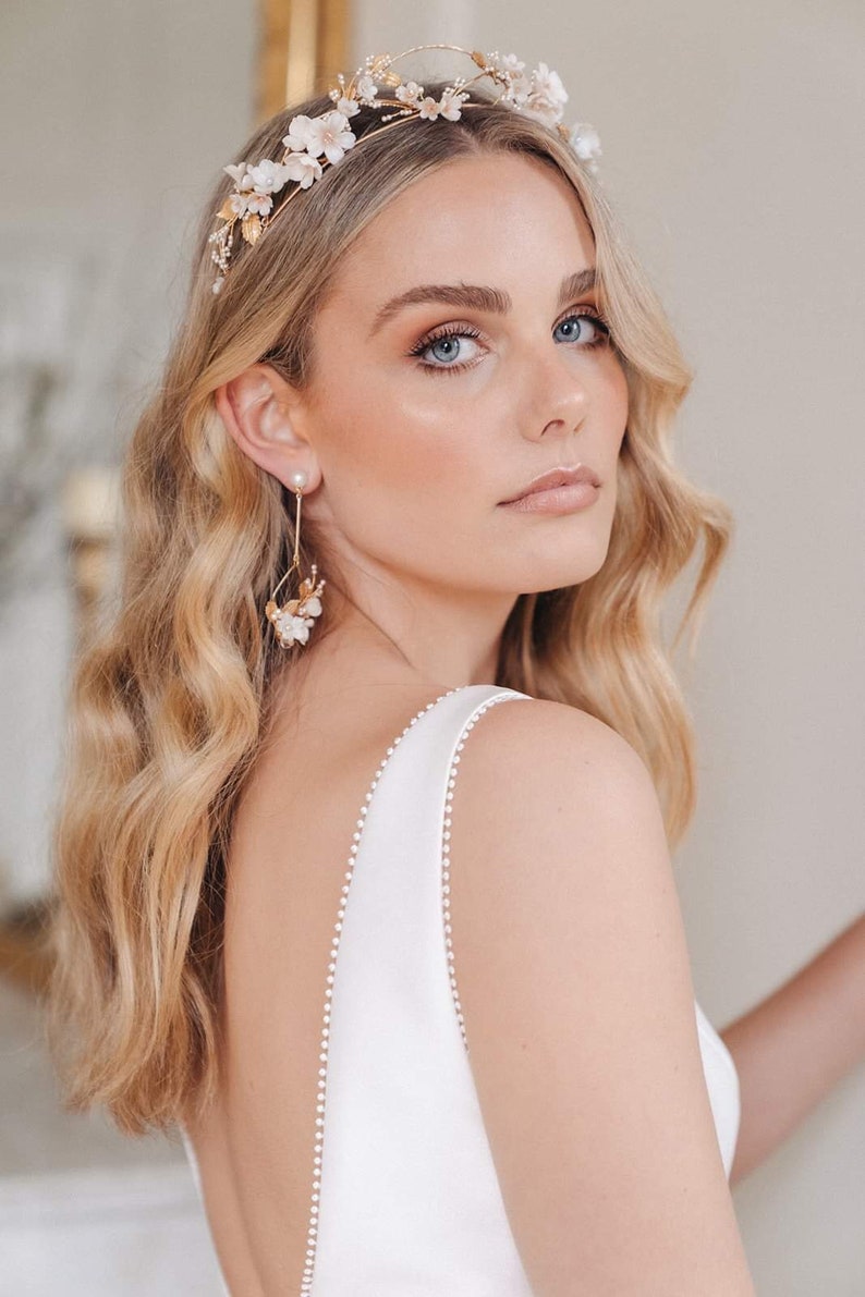 May include: A woman wearing a white dress with a delicate pearl trim, a gold floral crown, and matching gold floral earrings. The crown and earrings feature white flowers and pearls.