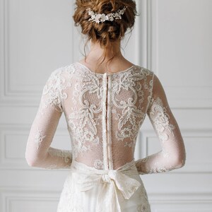 May include: A white lace wedding dress with long sleeves and a low back. The dress has a delicate floral pattern and a satin ribbon tied at the waist. The bride is wearing a floral hair comb.