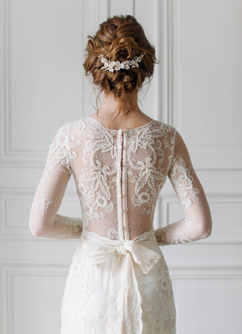 May include: A white lace wedding dress with long sleeves and a low back. The dress has a delicate floral pattern and a satin ribbon tied at the waist. The bride is wearing a floral hair comb.