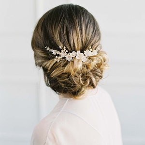 May include: A woman with brown hair styled in a low bun with a delicate floral hair comb. The comb features small white flowers and gold accents.