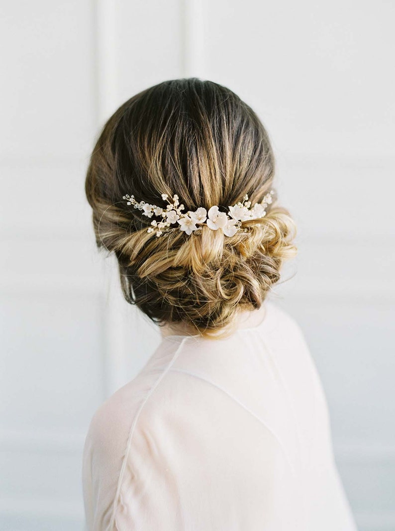 May include: A woman with brown hair styled in a low bun with a delicate floral hair comb. The comb features small white flowers and gold accents.