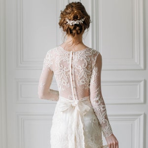 May include: A white lace wedding dress with a long sleeve and a bow at the waist. The dress has a lace pattern and a sheer back. The bride is wearing a floral hairpiece.