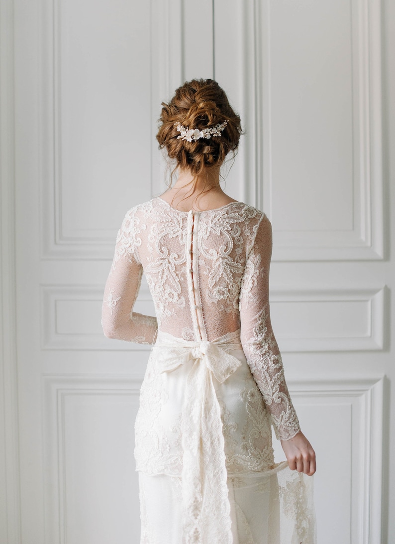May include: A white lace wedding dress with a long sleeve and a bow at the waist. The dress has a lace pattern and a sheer back. The bride is wearing a floral hairpiece.