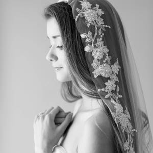 Puede incluir: Una novia con un vestido blanco sin tirantes y un velo de encaje delicado con adornos florales. El velo est&aacute; drapeado sobre su cabeza y hombros, enmarcando su rostro.