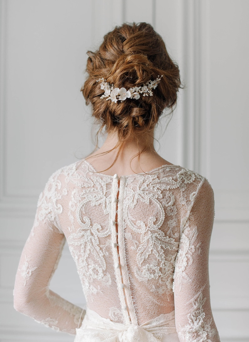 May include: A bride wearing a white lace wedding dress with a floral hairpiece. The dress has a low back with a row of buttons.