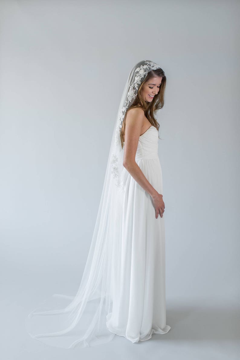 Puede incluir: Una novia con un vestido de novia blanco con un velo largo y fluido. El velo tiene un delicado encaje.