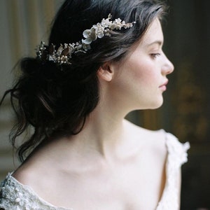 May include: A woman with dark hair wearing a white off-the-shoulder dress with floral embroidery. She is wearing a delicate floral hairpiece with pearl and gold accents.