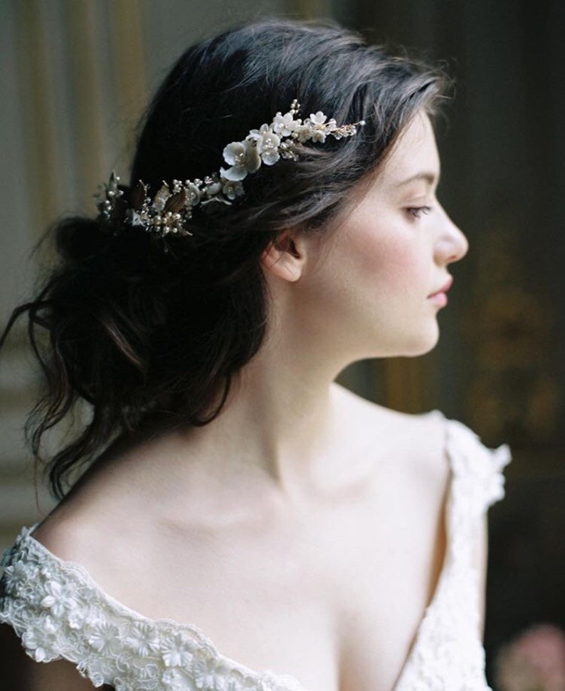 May include: A woman with dark hair wearing a white off-the-shoulder dress with floral embroidery. She is wearing a delicate floral hairpiece with pearl and gold accents.