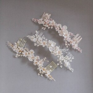 May include: Three hair combs with delicate floral designs. Each comb features a cluster of white and blush-colored flowers with pearl accents and crystal embellishments. The combs are arranged in a staggered pattern on a gray background.